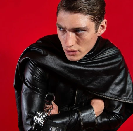 Adult male model in a black leather jacket posing in front of a red background, showcasing a modern and edgy fashion aesthetic.