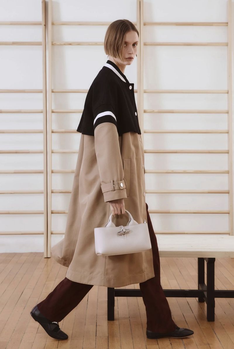 Fashion editorial shot of a woman walking with a handbag in a beige trench coat and burgundy pants in an indoor gym setting.
