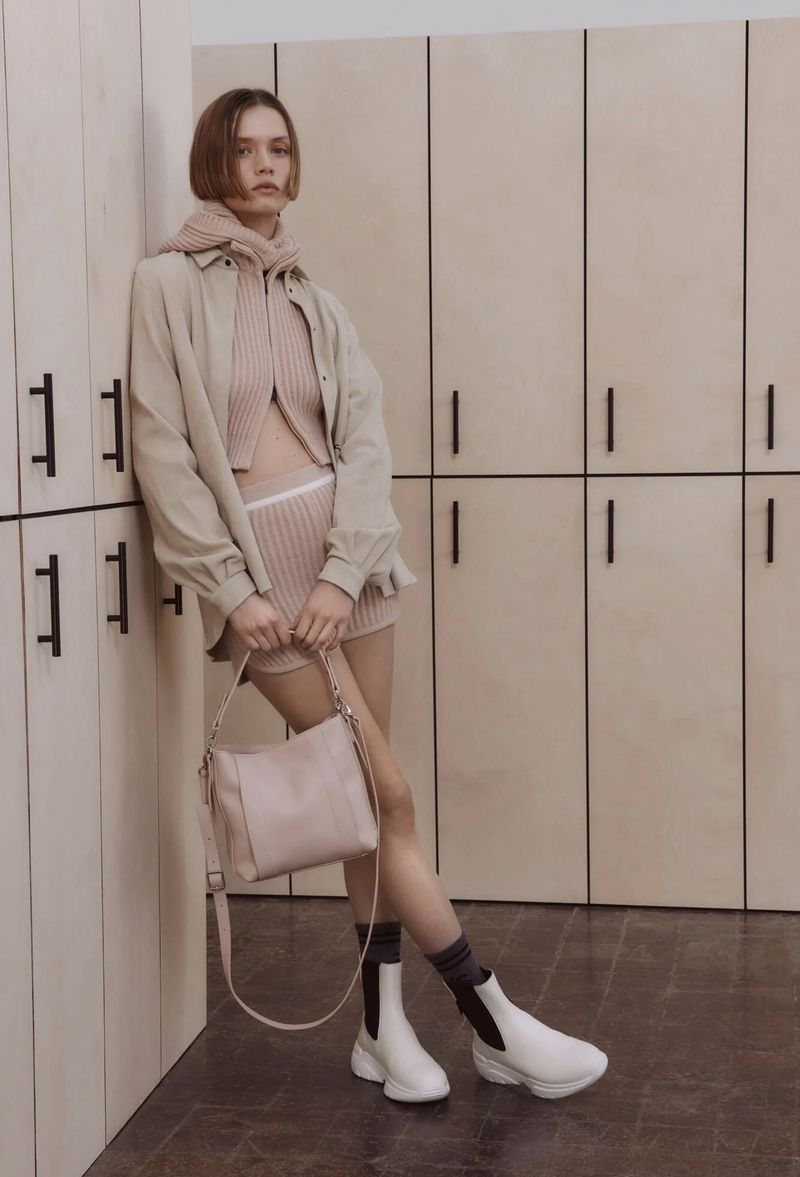 An adult woman posing in a sandy color coordinated outfit next to a wall of lockers indoors, holding a matching handbag and wearing boots.