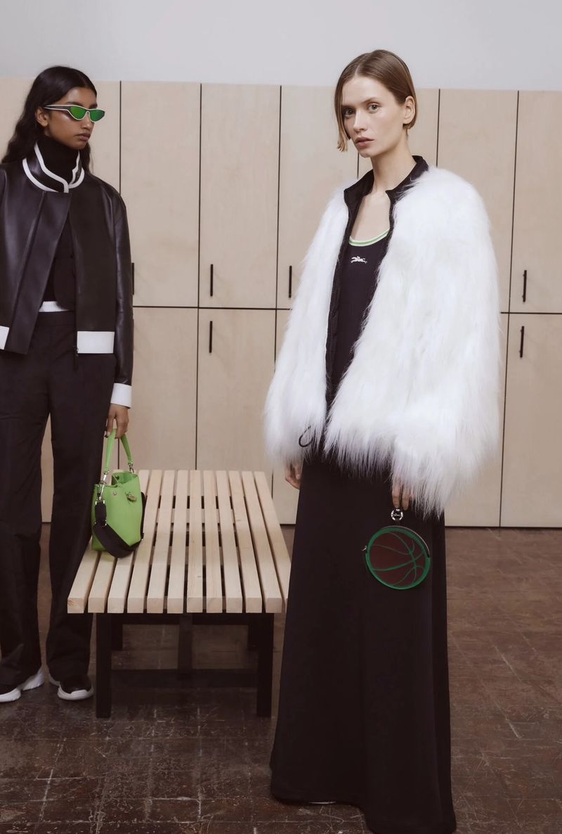 Two fashion models pose indoors against lockers; one wears a white faux fur coat, the other a black bomber jacket and green sunglasses.