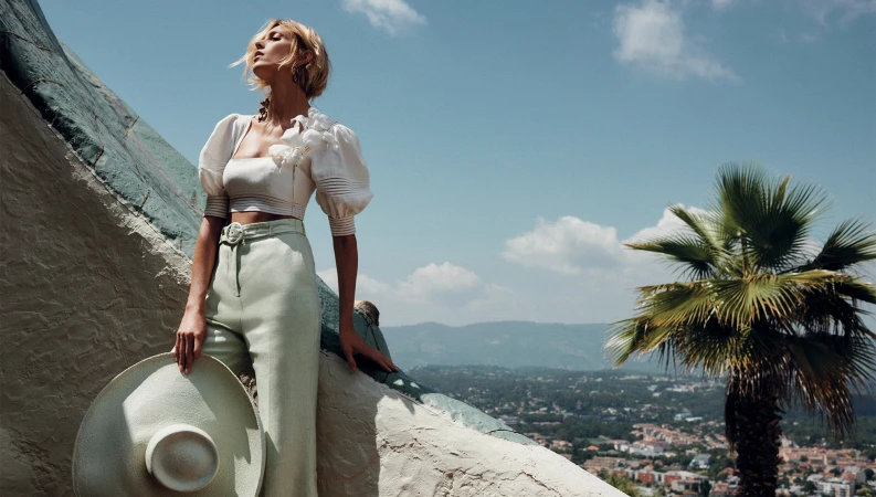 A fair-skinned woman in a light-colored linen outfit stands on a patterned wall, holding a hat, with a palm tree and city in the background.