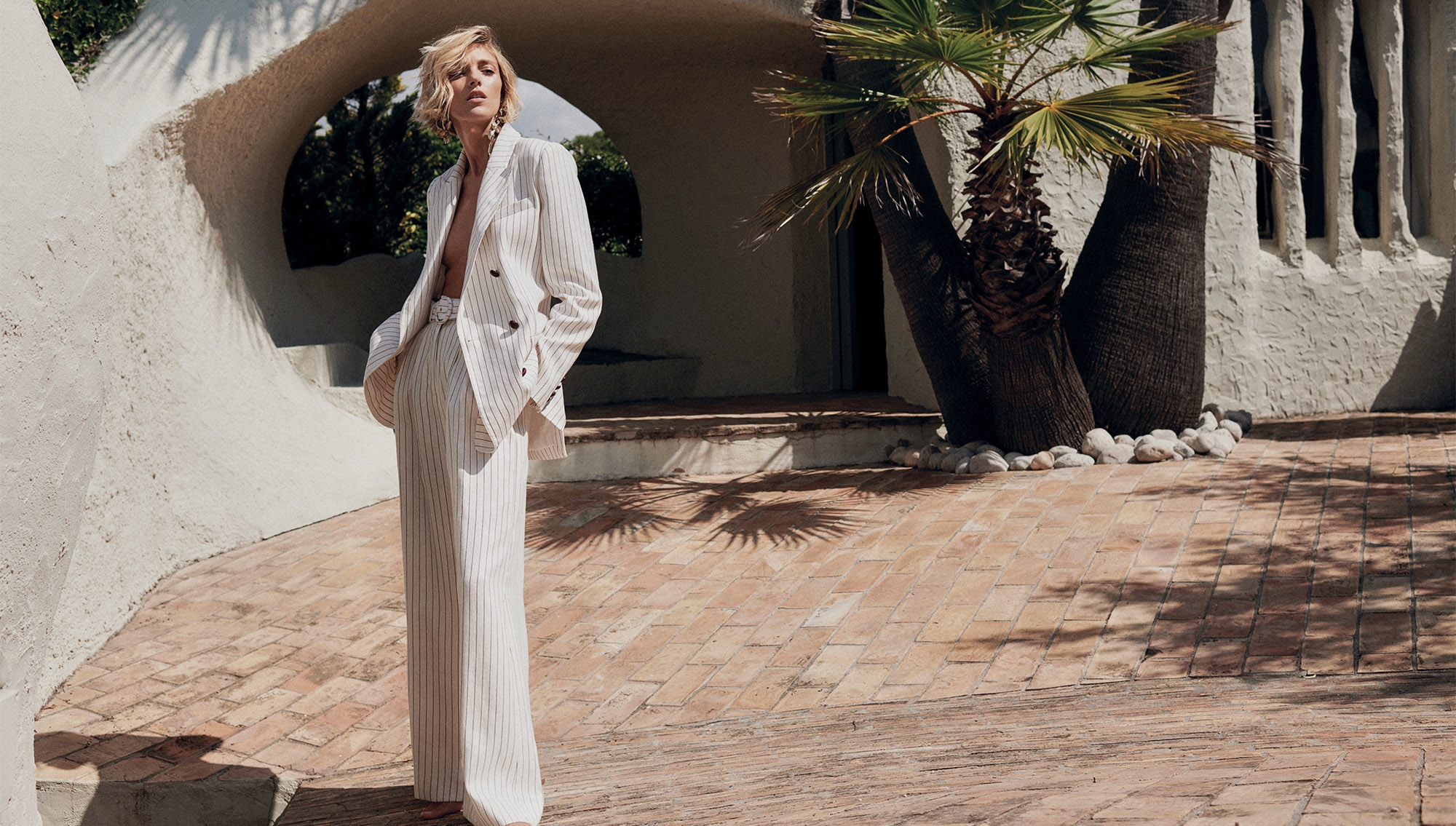Anja Rubik wearing a pinstripe pantsuit in front of a stucco building and palm tree