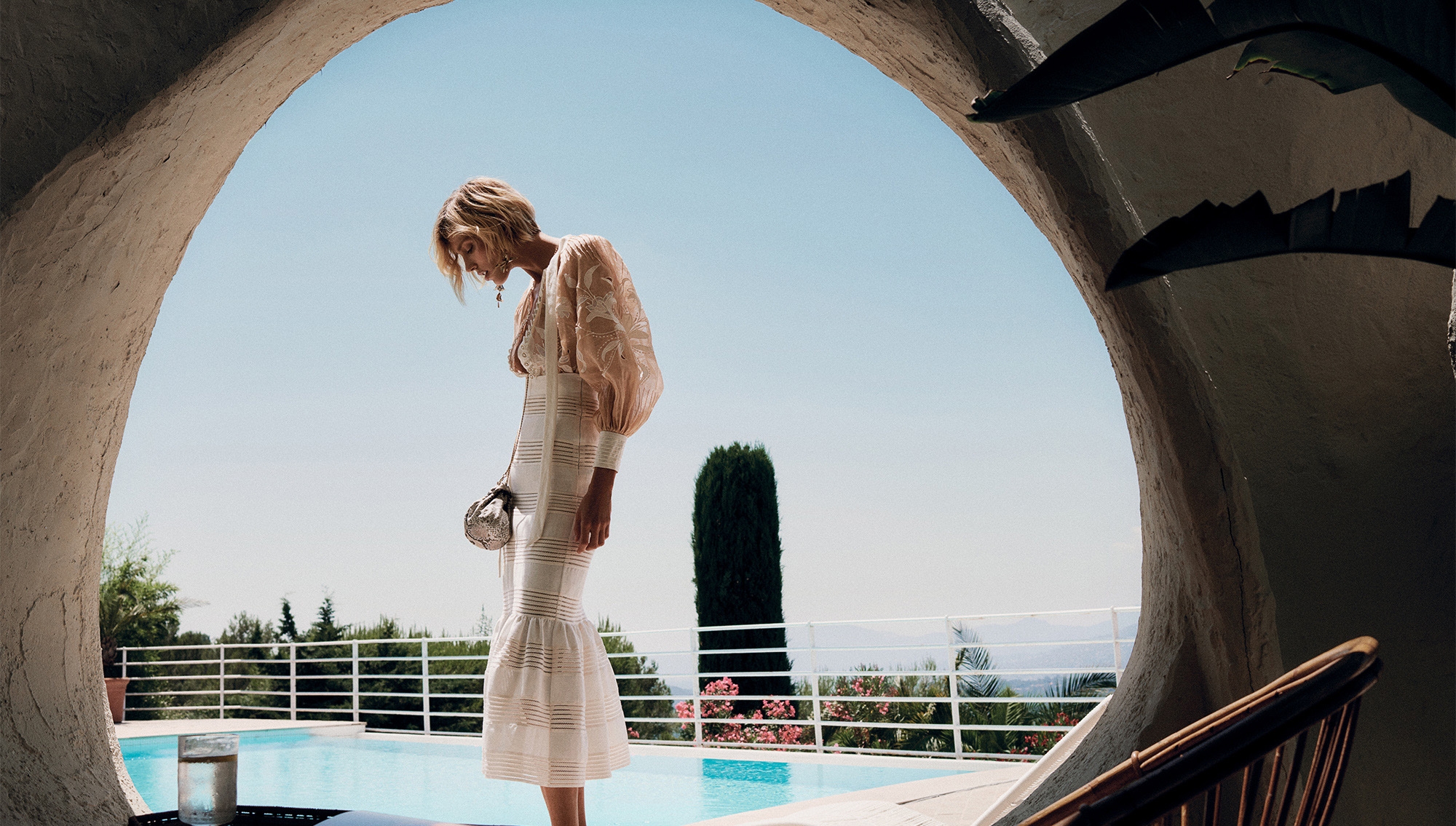 Woman in flowing dress stands by pool with blue sky background, framed by an arched doorway overlooking a sunny scene.