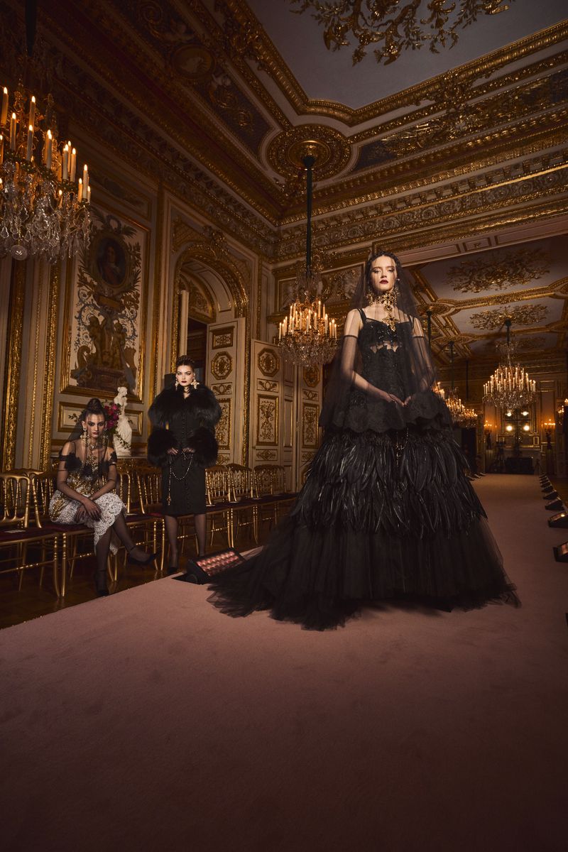 Runway scene with three women in elaborate black dresses, chandeliers, and ornate gold decor at fashion show inside a ballroom.