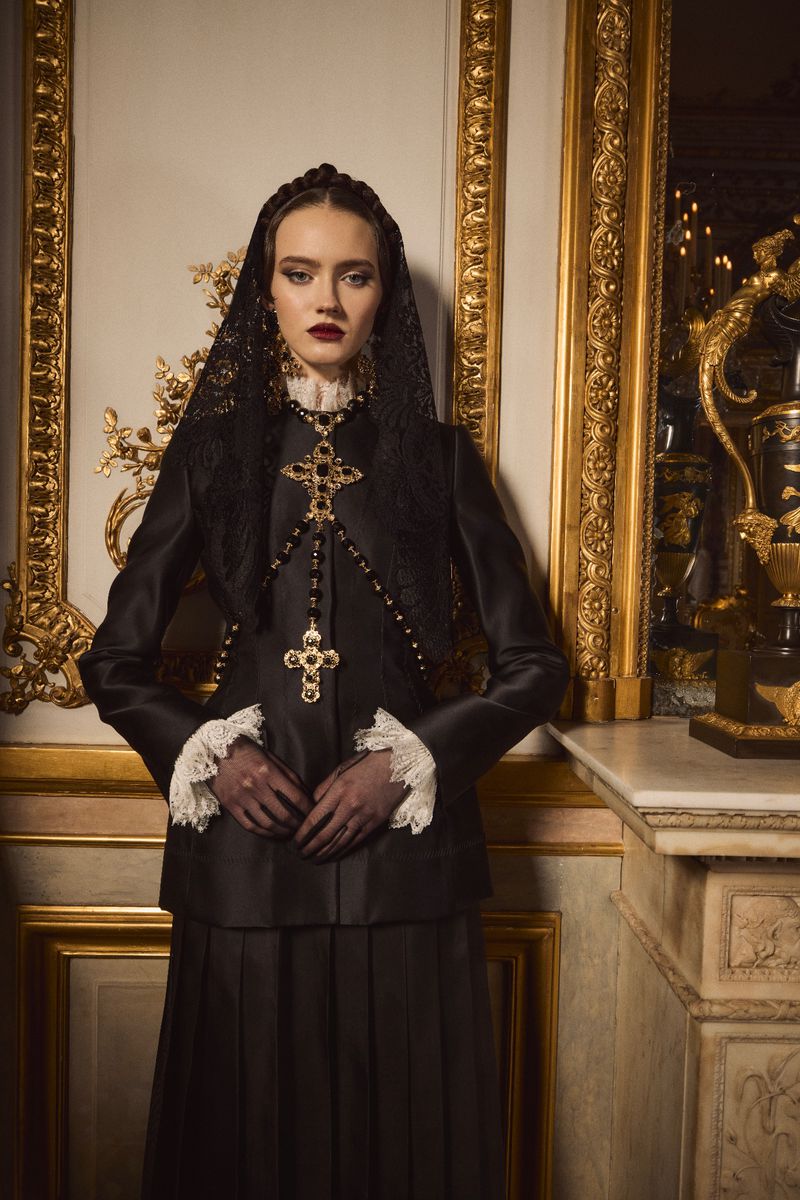 Fashionable woman in black dress, lace veil, and cross necklaces stands indoors against a gold frame, exuding elegance and style.
