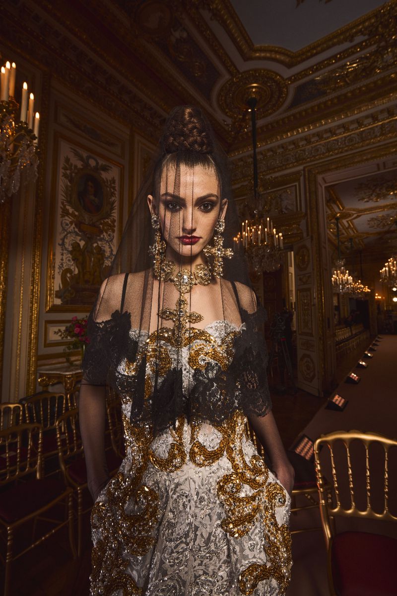 A model poses in an ornate gown with elaborate gold details, complemented by statement jewelry in a gilded hall with chandeliers.