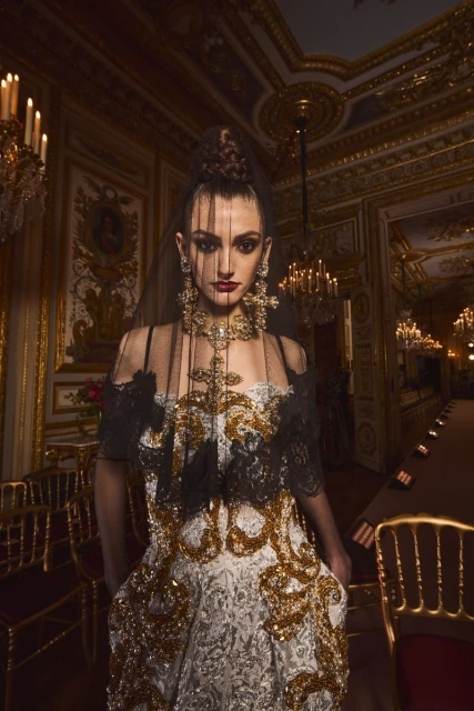 A model poses in an ornate gown with elaborate gold details, complemented by statement jewelry in a gilded hall with chandeliers.