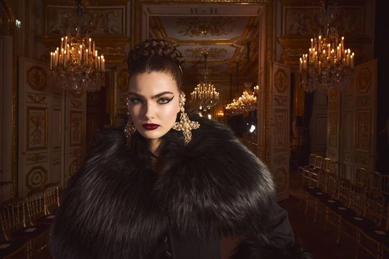 Fashionable woman in fur coat and large earrings poses in a grand hall with chandeliers, gold accents, and rows of chairs.