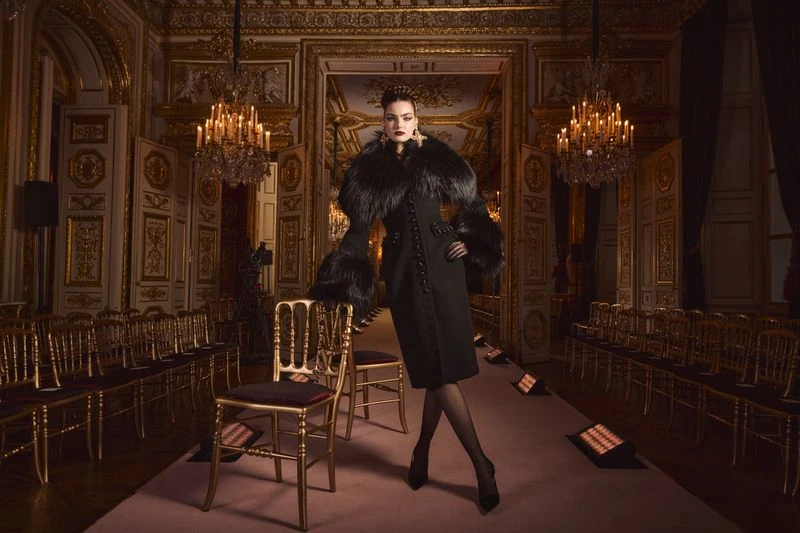 Woman in an elegant black coat with fur collar stands in a luxurious ballroom with golden chandeliers and chairs for a fashion show.