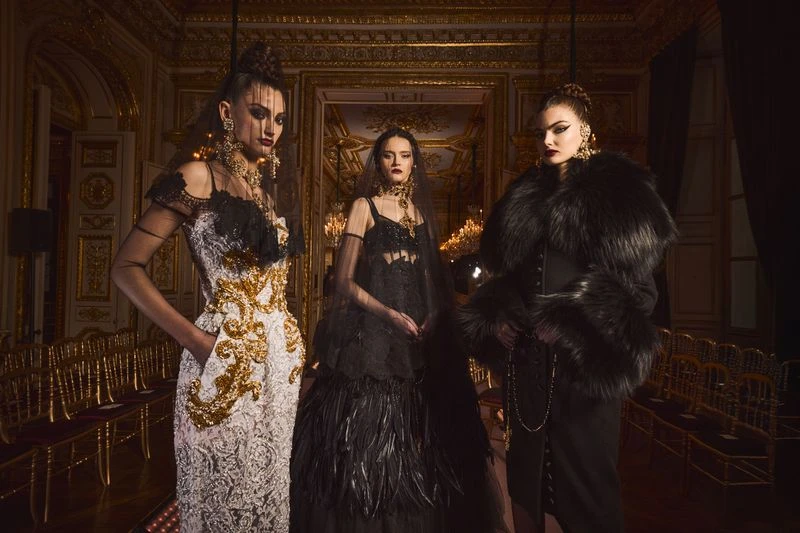 Three adult women in formal black-and-gold gowns pose in a ballroom with gold chandeliers, framed by gold chairs and decorative architecture.