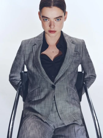 Model seated on a chair wearing a grey tailored suit jacket and black shirt against a plain white background