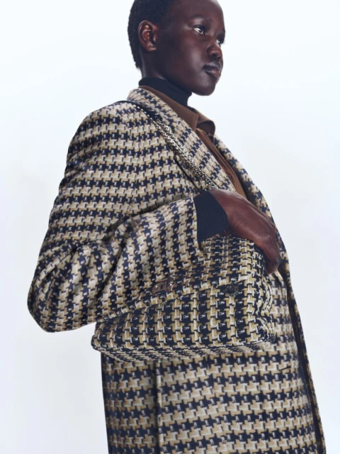 Fashionable woman wearing a houndstooth blazer and brown turtleneck against a white backdrop in a studio setting.