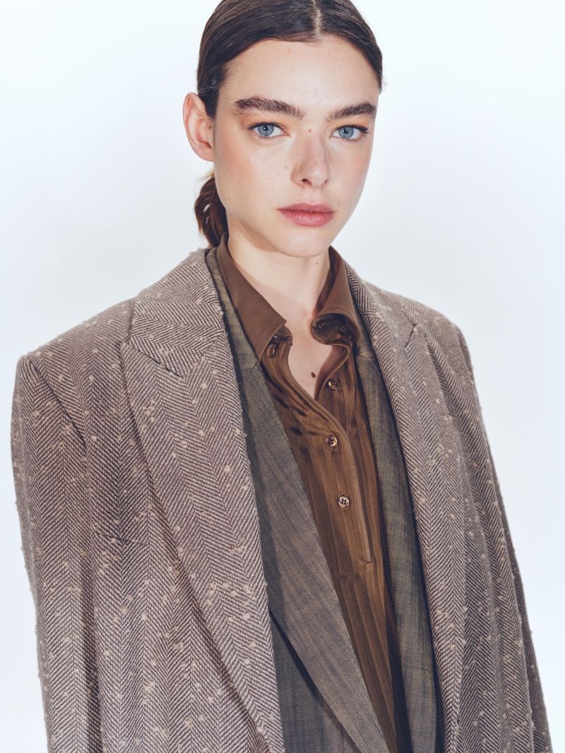 Headshot of an adult woman in a brown jacket and shirt against a white backdrop, looking directly at the camera with a neutral expression.