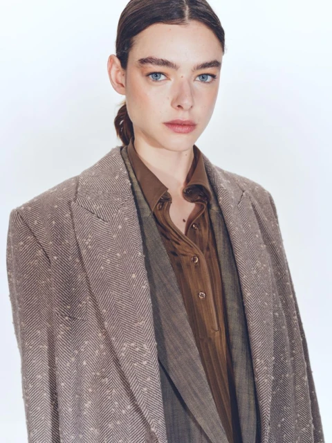 Headshot of an adult woman in a brown jacket and shirt against a white backdrop, looking directly at the camera with a neutral expression.