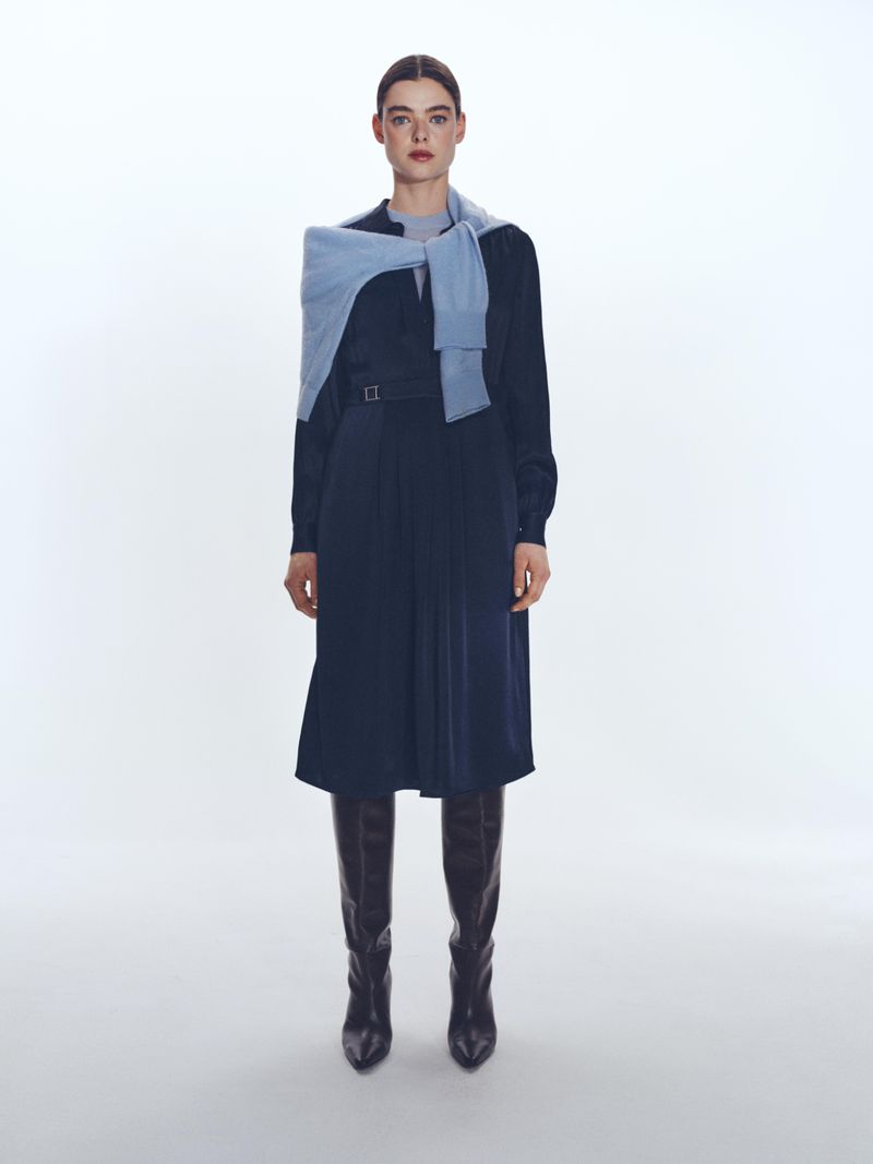 Model standing against a white seamless background wearing a navy midi dress with a light blue sweater draped over shoulders and dark boots