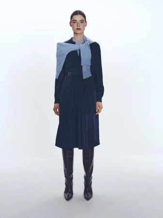 Model standing against a white seamless background wearing a navy midi dress with a light blue sweater draped over shoulders and dark boots