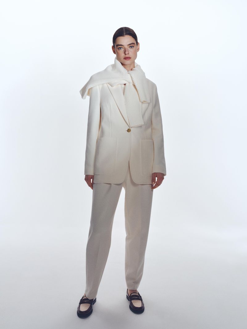 Full-shot fashion portrait of a woman in a cream-colored pantsuit and black loafers, posing against a bright white backdrop with soft studio lighting.