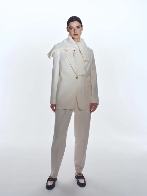 Full-shot fashion portrait of a woman in a cream-colored pantsuit and black loafers, posing against a bright white backdrop with soft studio lighting.