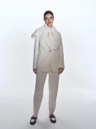 Full-shot fashion portrait of a woman in a cream-colored pantsuit and black loafers, posing against a bright white backdrop with soft studio lighting.