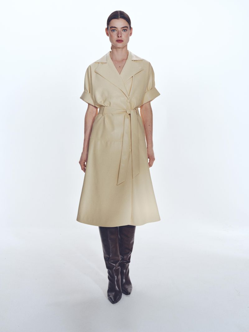 Full-length shot of a woman in a beige trench coat dress and black leather boots against a bright white backdrop, fashion forward.