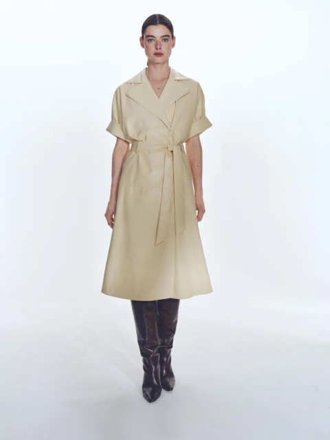 Full-length shot of a woman in a beige trench coat dress and black leather boots against a bright white backdrop, fashion forward.