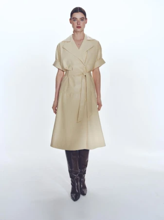 Full-length shot of a woman in a beige trench coat dress and black leather boots against a bright white backdrop, fashion forward.