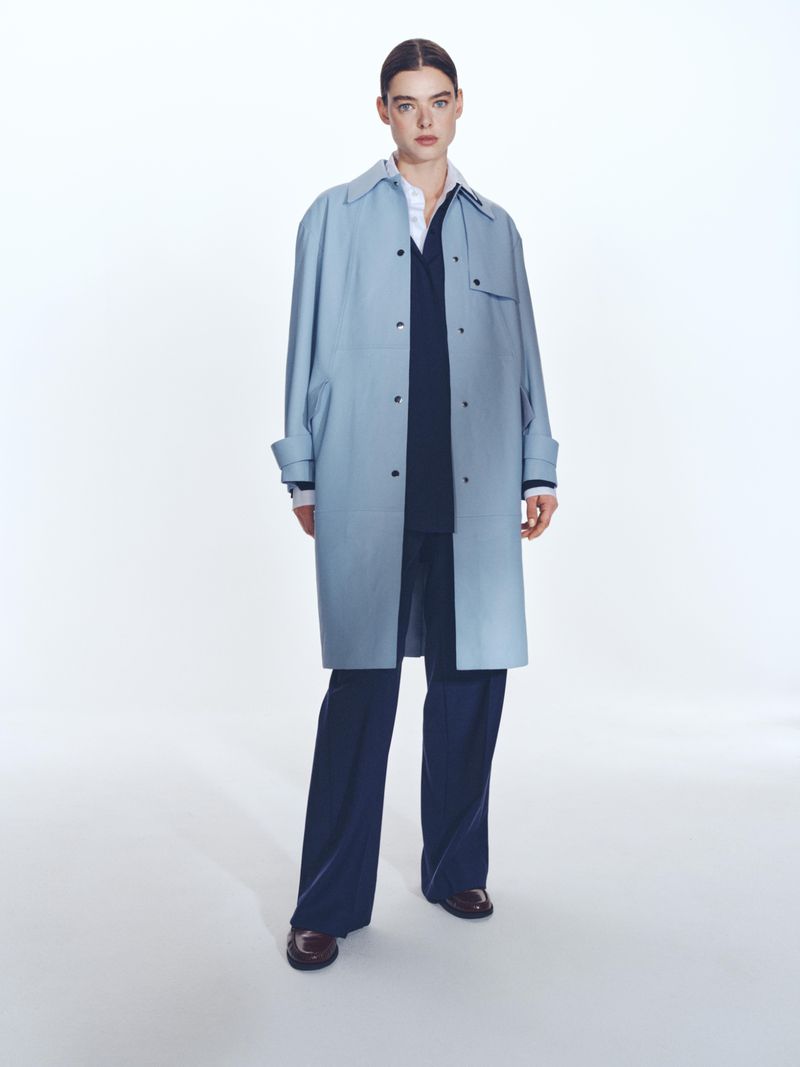 A woman models a light blue trench coat, layered over a navy outfit and paired with maroon loafers, in a bright, full-length studio shot.