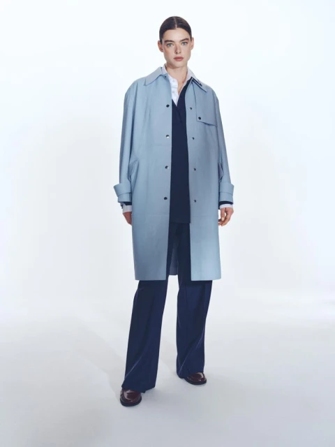 A woman models a light blue trench coat, layered over a navy outfit and paired with maroon loafers, in a bright, full-length studio shot.