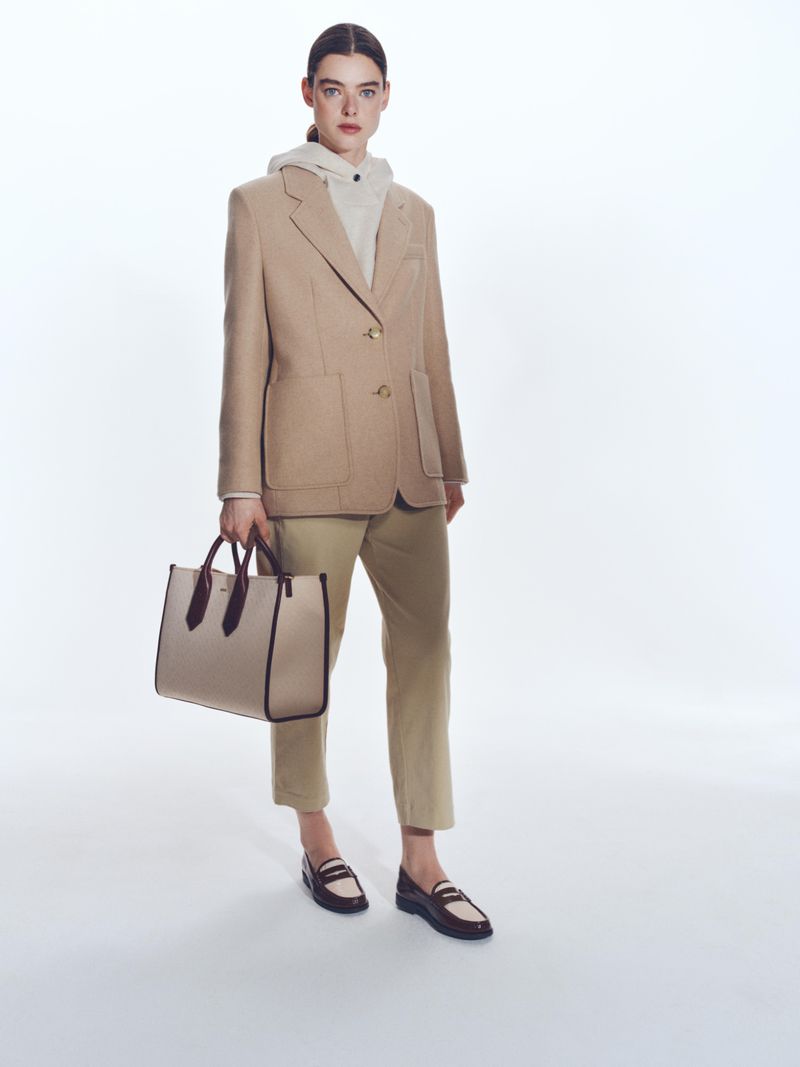 Fashionable adult female stands wearing blazer, hoodie, trousers, and loafers, holding a handbag against a white backdrop.