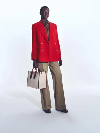 Model wearing a bright red blazer, brown trousers, and holding a structured tote bag against a white studio background.