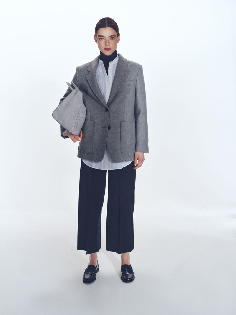 Elegant woman in a grey blazer, striped shirt, and black culottes, holding a grey tote bag, standing in a bright white photo studio.