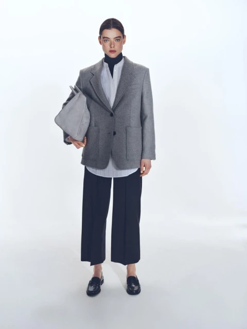 Elegant woman in a grey blazer, striped shirt, and black culottes, holding a grey tote bag, standing in a bright white photo studio.