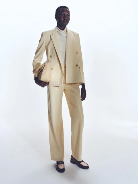Fashion shot of an African American adult female wearing a light yellow pantsuit and holding a bag, studio setting with a white background.