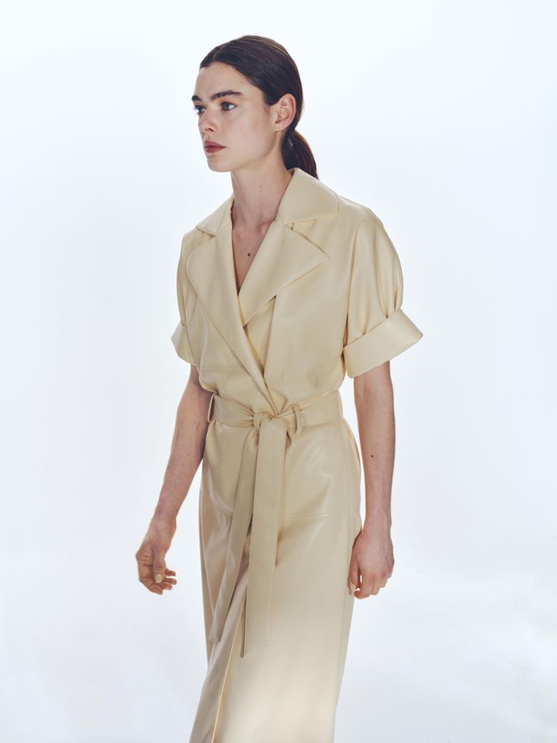 Elegant woman in a cream-colored wrap dress poses in a bright studio. The portrait showcases a modern and minimalist fashion aesthetic.