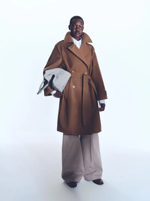 An adult woman models a full-length belted trench coat with wide leg pants, holding a bag, in a studio setting on a white background.