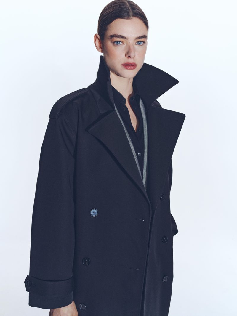 Model wearing a dark double-breasted trench coat with a popped collar against a plain white studio background