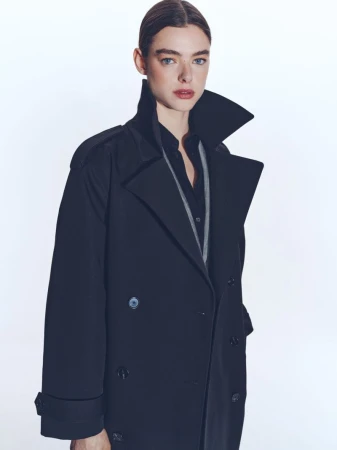 Model wearing a dark double-breasted trench coat with a popped collar against a plain white studio background