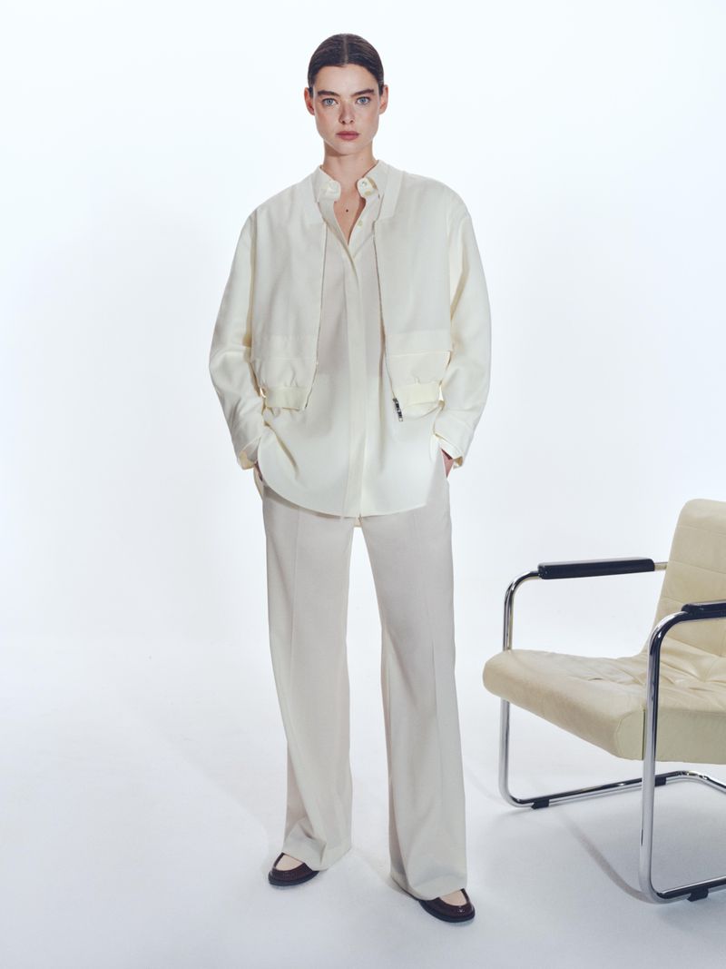 Studio shot of a young woman in a casual cream-colored matching set, standing indoors next to an off-white lounge chair.