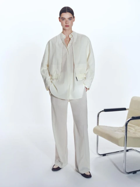 Studio shot of a young woman in a casual cream-colored matching set, standing indoors next to an off-white lounge chair.