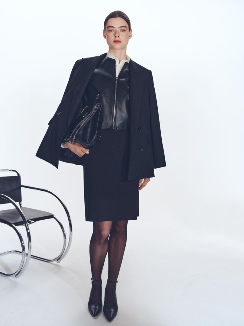 Model standing in a black blazer, leather top, and skirt holding a large black handbag against a white studio background.