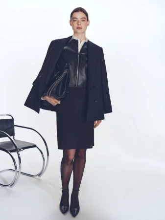 Model standing in a black blazer, leather top, and skirt holding a large black handbag against a white studio background.
