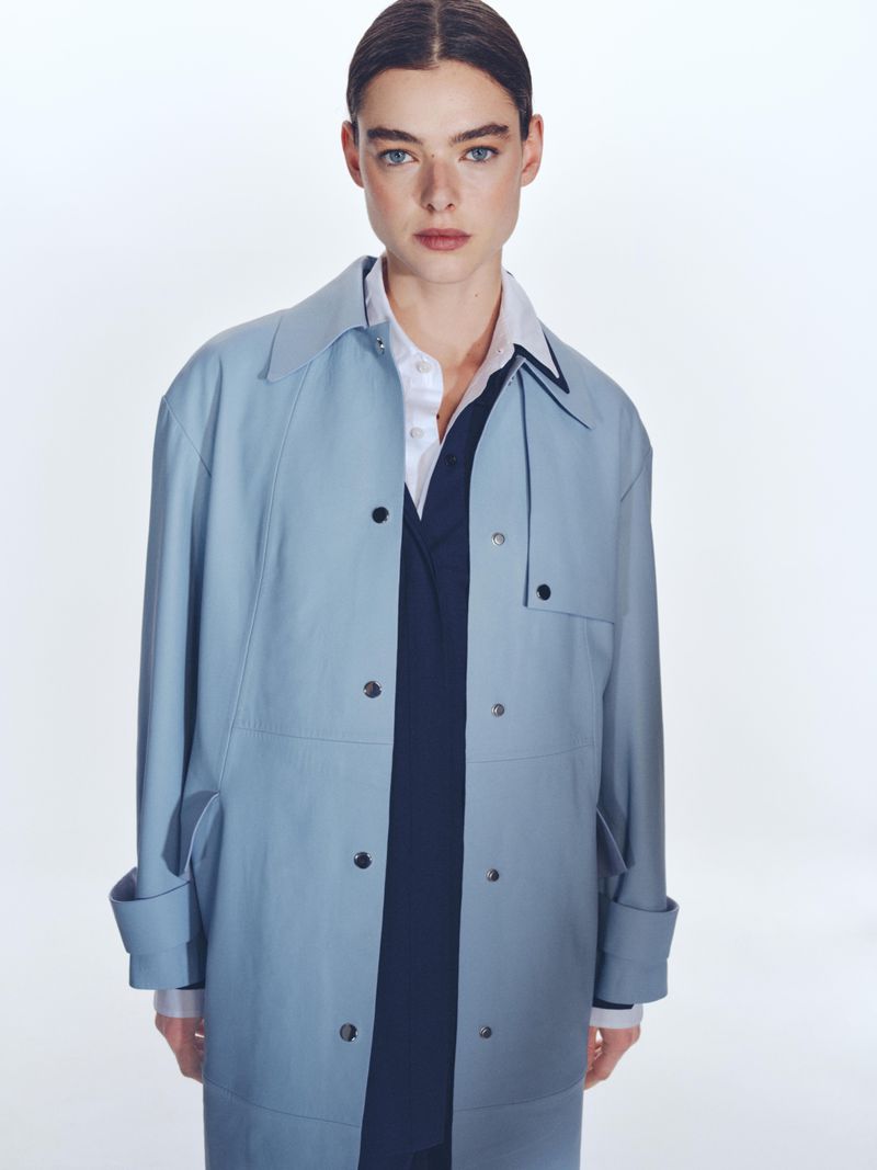 Portrait of a stylish adult female wearing a light blue trench coat, white button-down shirt, and navy v-neck sweater against a white backdrop.