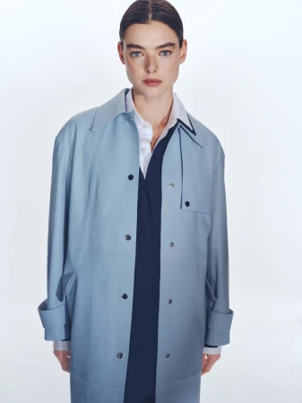 Portrait of a stylish adult female wearing a light blue trench coat, white button-down shirt, and navy v-neck sweater against a white backdrop.