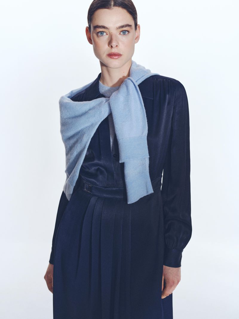 Model in dark blue dress wearing a light blue sweater tied around shoulders against a white background