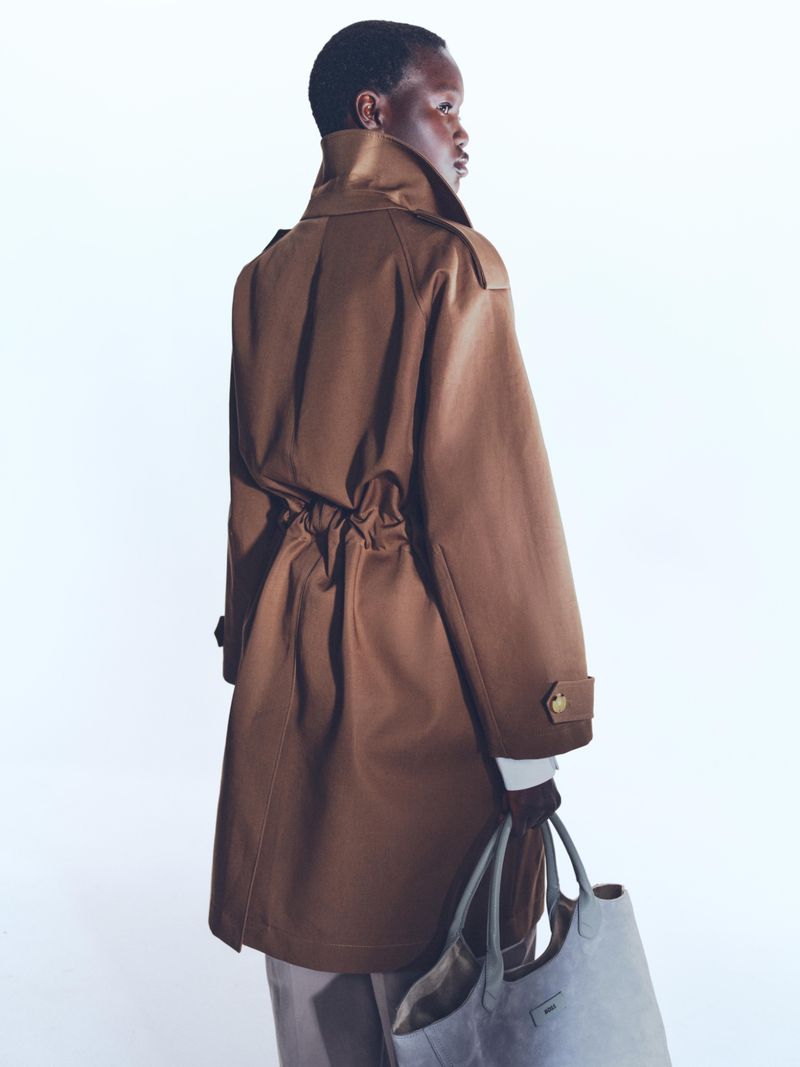 Model wearing a brown belted coat and holding a light grey tote bag against a bright white background