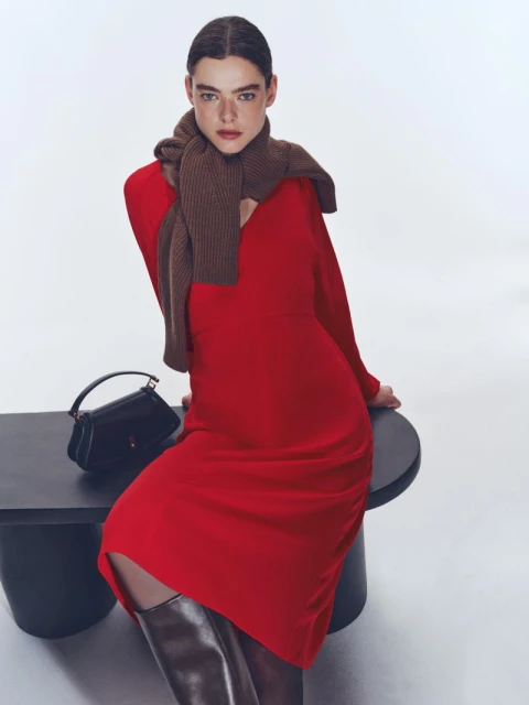 Brunette woman in red long-sleeved dress and brown scarf poses seated on a black table with a black purse against a white backdrop.