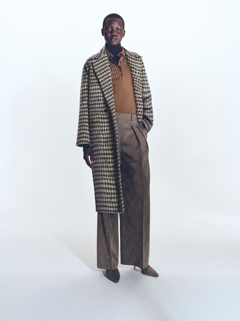 A full-length shot of a person in a patterned coat and wide-leg trousers stands against a clean white backdrop in a fashion editorial.