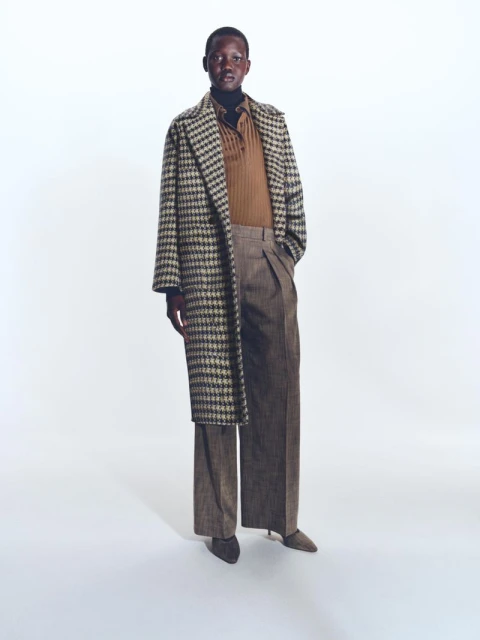 A full-length shot of a person in a patterned coat and wide-leg trousers stands against a clean white backdrop in a fashion editorial.