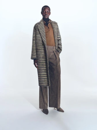 A full-length shot of a person in a patterned coat and wide-leg trousers stands against a clean white backdrop in a fashion editorial.