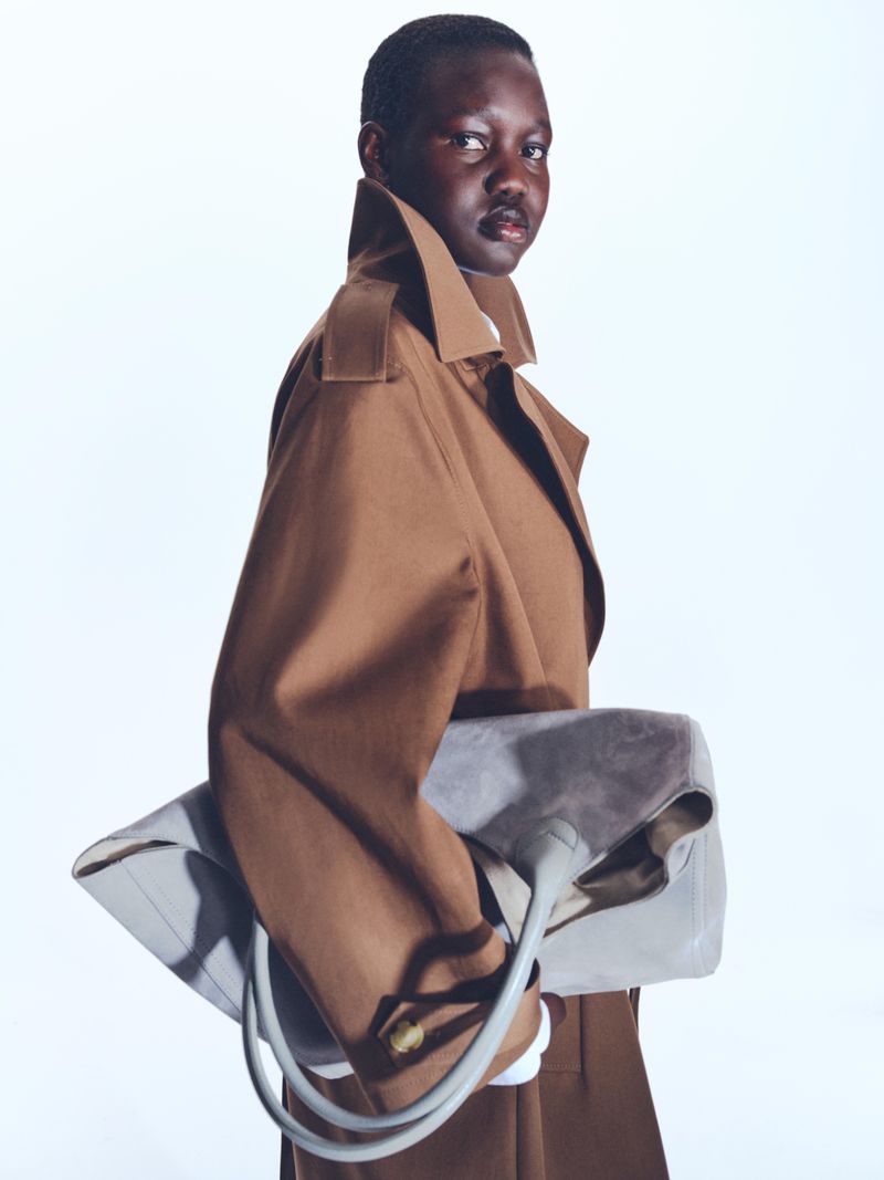 Stylish woman in a tan trench coat holds a grey leather handbag in a fashion studio with a bright white background.